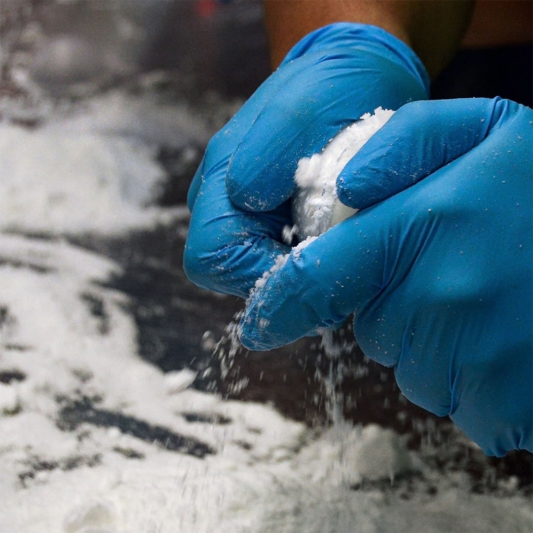 Close-up of gloved hands shaping a white Muscle Rehab bath bomb during manufacturing, with powdery ingredients scattered on the work surface, highlighting the handmade production process.