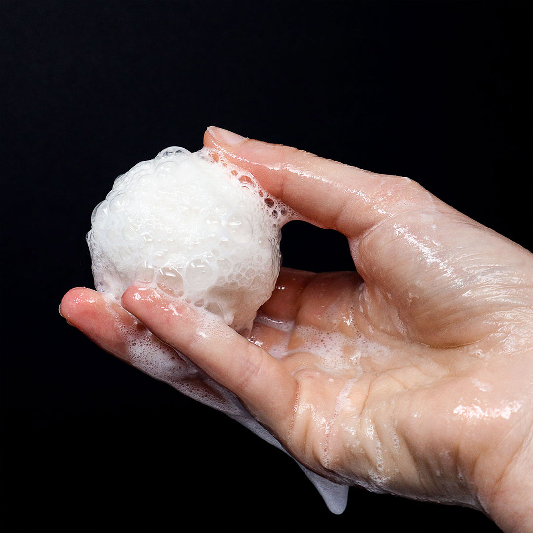 Close-up of a hand holding a fizzing white Muscle Rehab bath bomb covered in foamy bubbles against a black background, capturing the texture and effervescent action during use.