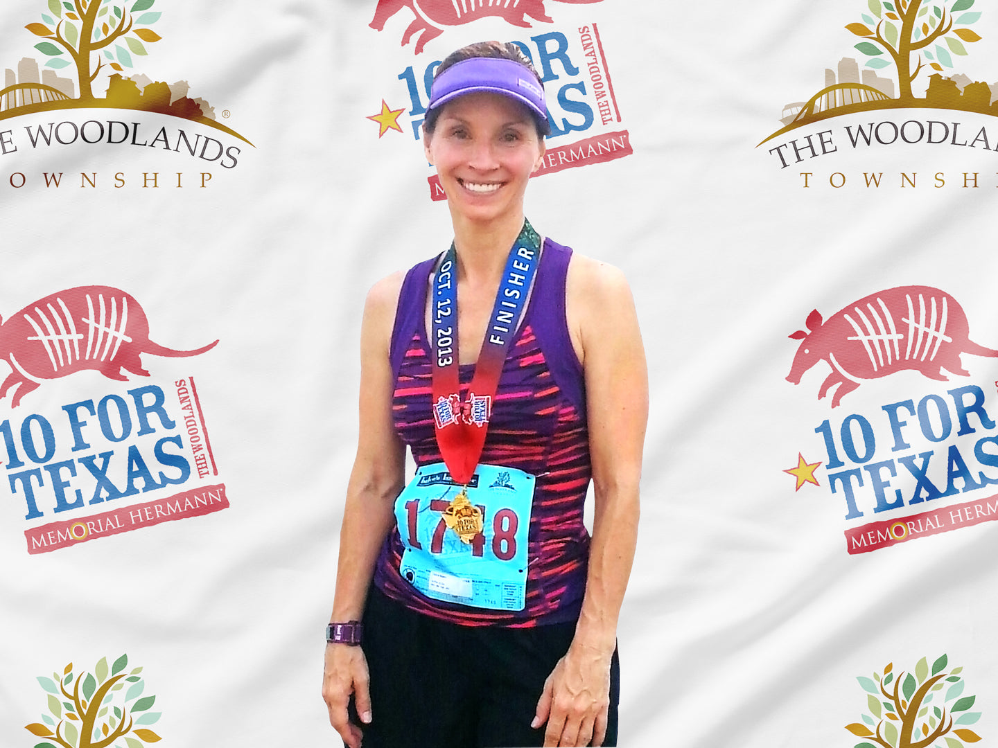 Debra Myers smiling and wearing a finisher’s medal and race bib after completing the “10 for Texas” event, standing in front of a backdrop featuring The Woodlands Township and event logos.