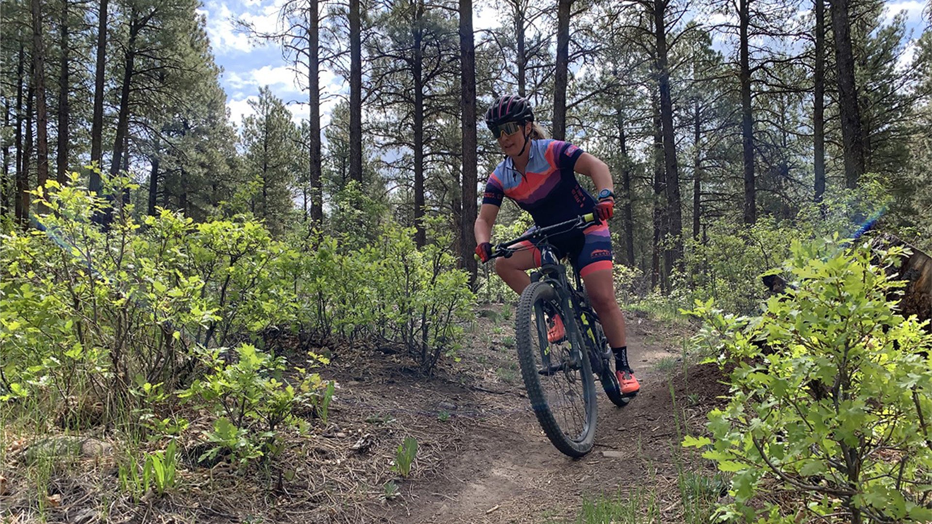 This image shows a cyclist riding a mountain bike down a forest trail. She is wearing a colorful cycling kit in shades of pink, purple, and navy, along with a black and red helmet, sunglasses, and bright orange shoes. The trail is surrounded by green bushes and tall pine trees, with sunlight filtering through, suggesting an active, adventurous outdoor setting.