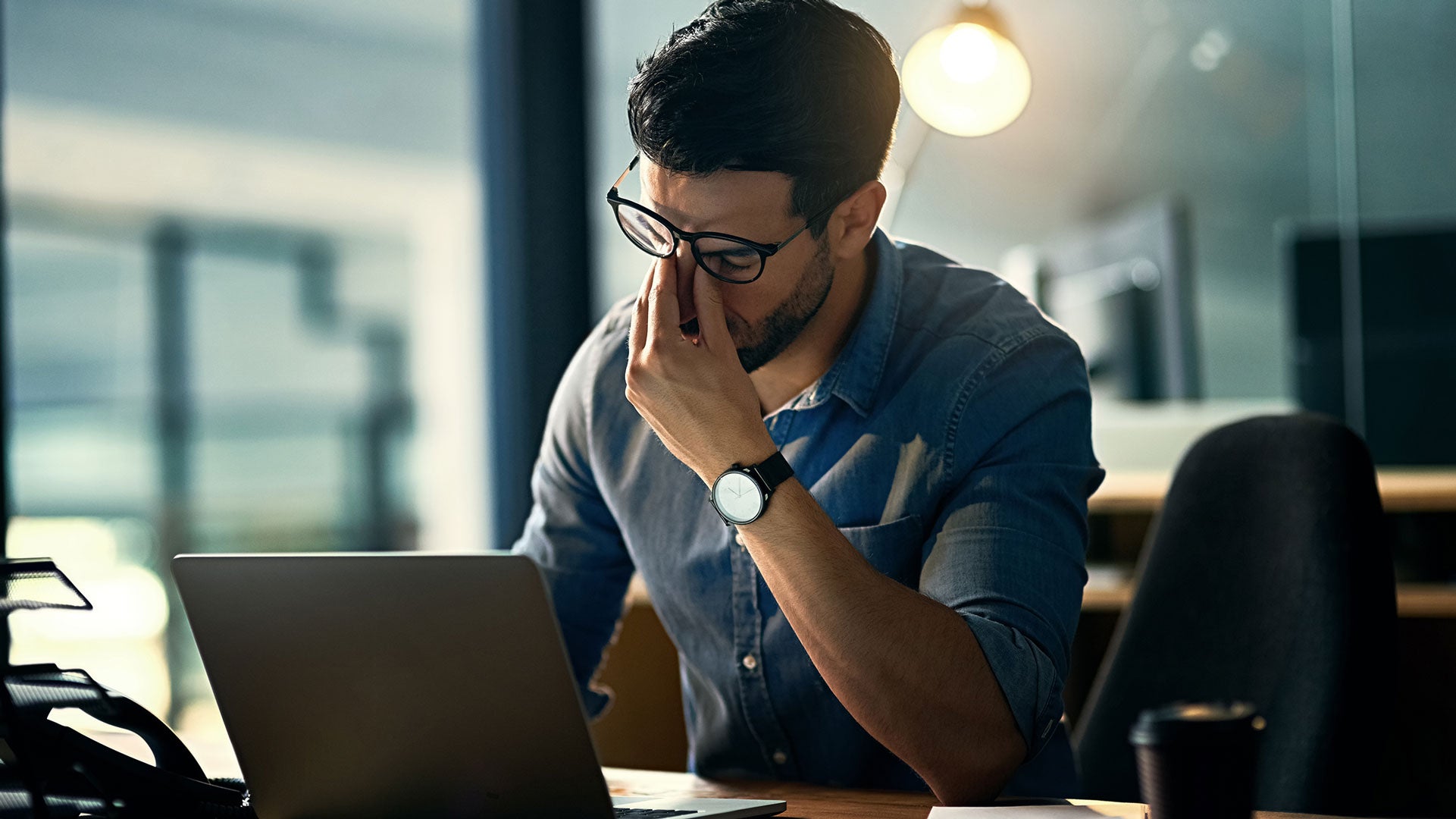 Man sitting at a desk with a laptop, holding his head in his hand, possibly indicating stress or exhaustion.