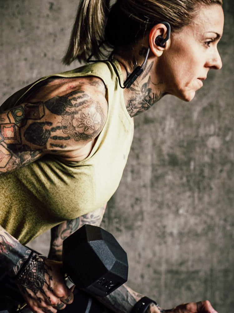 This image shows a focused, athletic woman (Carina Reid) performing a bent-over dumbbell row. She is wearing a sleeveless yellow workout top, black leggings, and wireless earphones. Her hair is tied back in a ponytail, and her arms, shoulders, neck, and hands are heavily tattooed with floral and geometric designs. The background is a textured concrete wall, giving the photo a gritty, high-intensity gym atmosphere.
