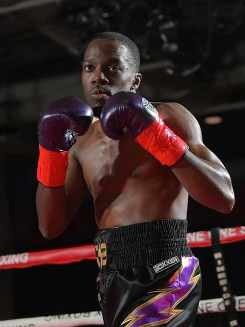 The image shows a male boxer (Joe Britton) in the ring, wearing purple boxing gloves, red wrist wraps, and black boxing shorts with purple and yellow lightning bolt designs. He is in a fighting stance, focused, and ready to throw or block punches, with the ropes of the boxing ring visible in the background.