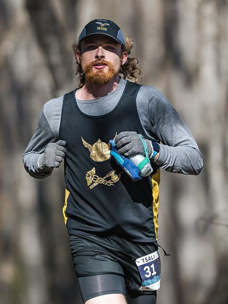 This image shows a man (Seth Hardwick) running outdoors in a race, wearing a black tank top with gold graphics over a long-sleeve gray shirt. He has a red beard, long hair pulled back, and is wearing a dark cap with the word "VESPA" on it. He’s holding a blue water bottle in one hand and wearing race bib number 31, indicating he’s participating in an organized running event.