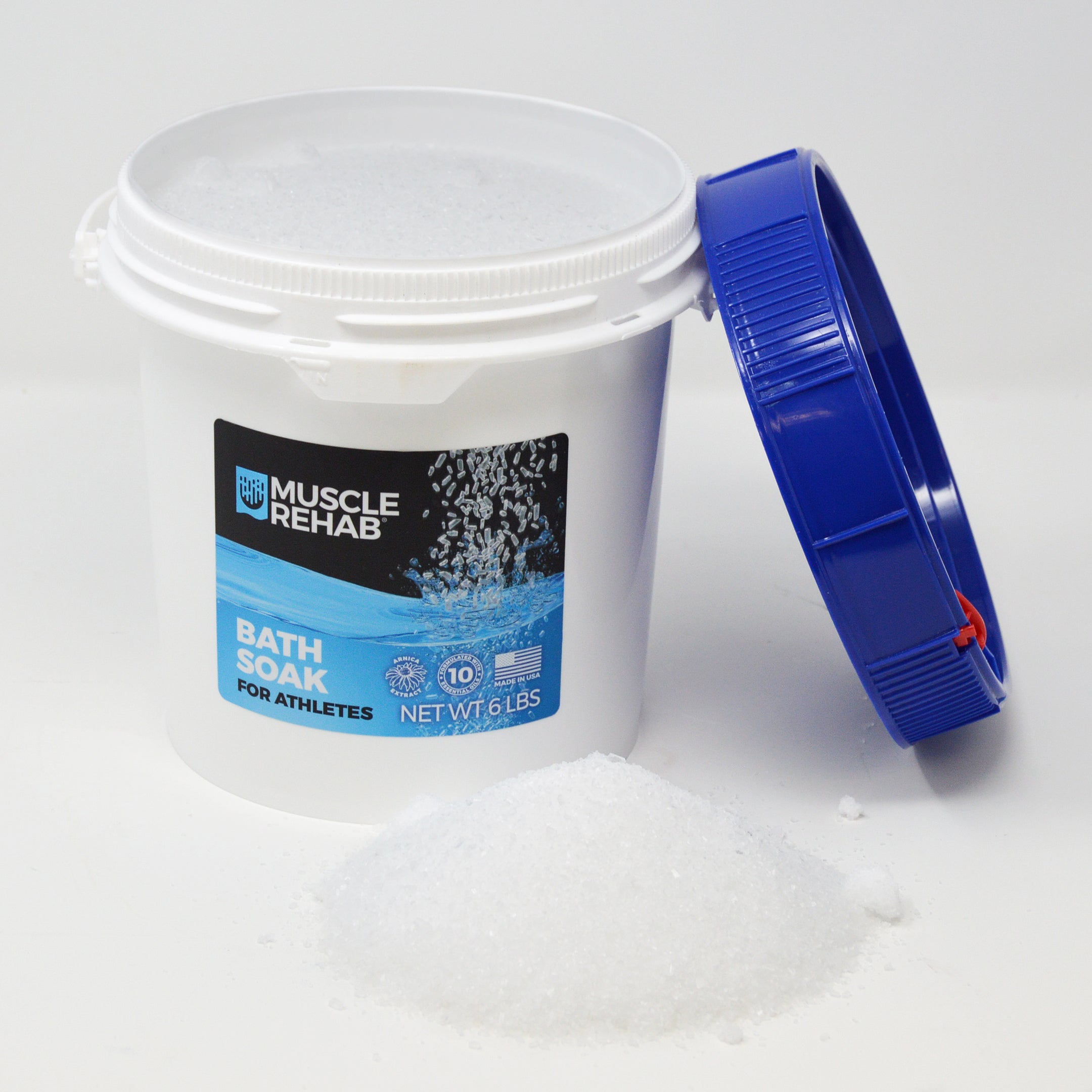 This image shows the Muscle Rehab® Bath Soak (6 lb pail) in a lifestyle/product display:
Product: White pail with a blue lid (lid is removed and placed to the side), showing the front label clearly.
Label Details: Brand logo, “Bath Soak for Athletes” name, “Formulated with 10 Essential Oils” badge, “Made in USA,” and net weight.
Lifestyle Element: A small mound of bath soak granules is poured in front of the container, showing the texture and crystalline appearance of the product.