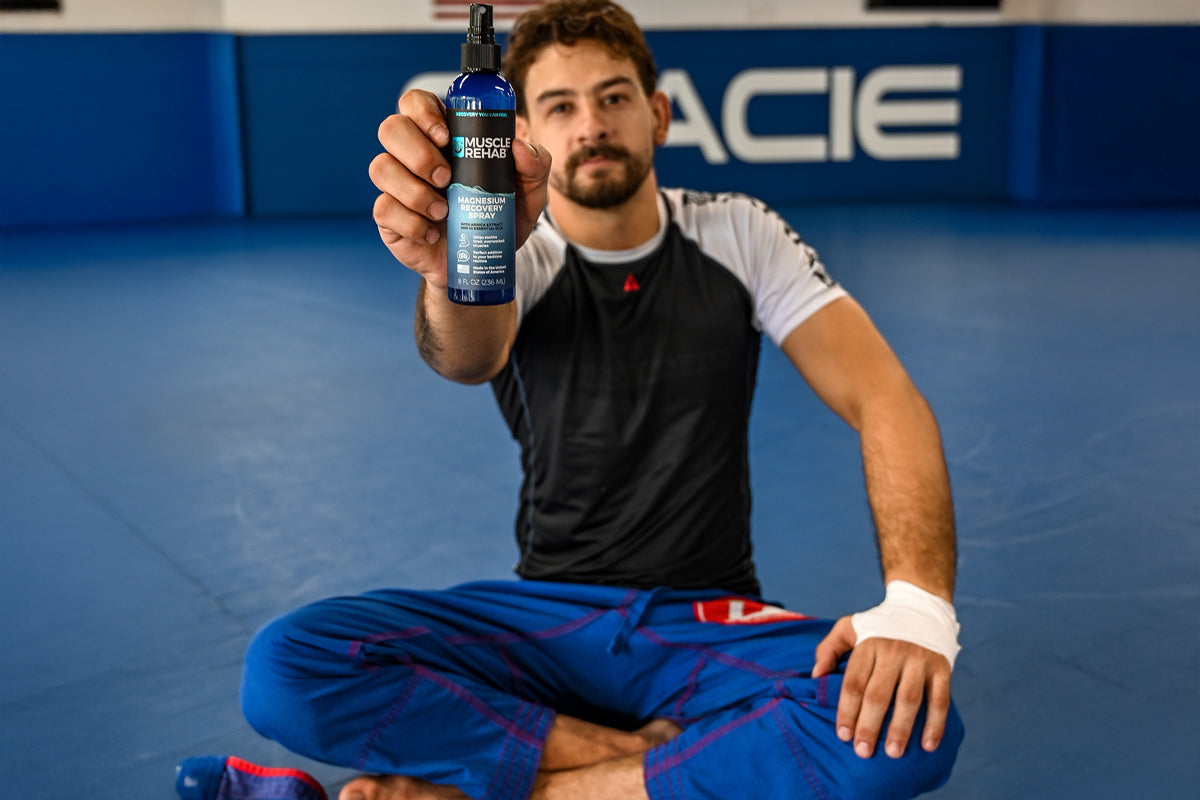 Athlete holding Muscle Rehab Magnesium Recovery Spray bottle while seated on a gym mat.