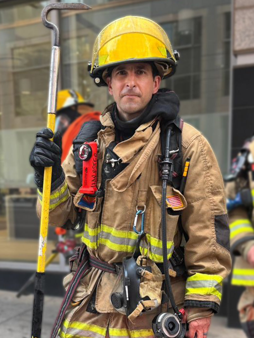 The image shows a firefighter (Nathan Harpster) in full turnout gear, including a yellow helmet, tan protective suit with reflective stripes, gloves, and breathing apparatus. He is holding a Halligan tool and standing in what appears to be an urban environment with other firefighters visible in the background.